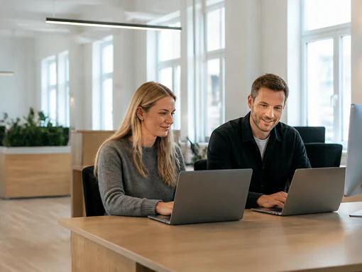 To kolleger sidder ved samme bord med hver sin bærbare computer på et lyst kontor